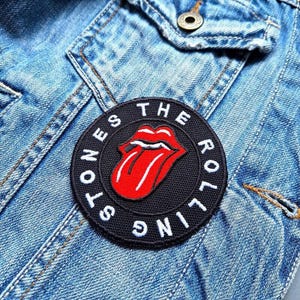 May include: A black circular patch featuring the Rolling Stones' iconic red tongue and lips logo, with the band's name in white lettering around the edge. The patch is sewn onto a light blue denim jacket.