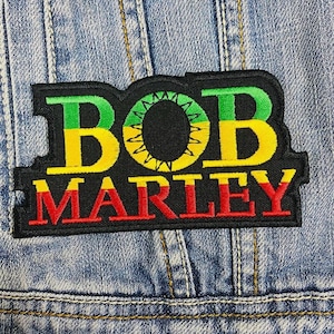May include: A black patch with the name "BOB MARLEY" in green, yellow, and red lettering. The patch is rectangular with a black border and is sewn onto a light blue denim jacket.