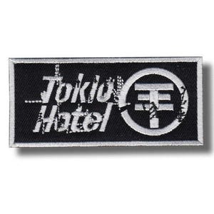 Può includere: Toppa rettangolare nera con testo ricamato bianco e un simbolo. Il testo recita "Tokio Hotel" in un carattere stilizzato. Un simbolo circolare con una linea orizzontale e verticale si trova sul lato destro.