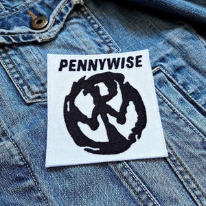 Pode incluir: Um patch quadrado branco com a palavra "PENNYWISE" em letras pretas sobre um desenho circular preto. O patch está em uma jaqueta jeans azul.