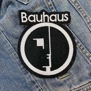 Könnte beinhalten: Ein schwarz-weißer Bauhaus-Aufnäher. Der Aufnäher zeigt das Wort "Bauhaus" in weißen Blockbuchstaben über einem schwarzen Kreis mit weißem Umriss. Im Inneren des Kreises befindet sich ein weißes geometrisches Design. Der Aufnäher befindet sich auf einer Jeansjacke.