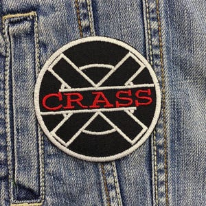 Peut inclure: Un écusson rond noir avec le mot "CRASS" en rouge. Le patch a une bordure blanche et un motif géométrique blanc. L'écusson est cousu sur une veste en jean bleue.