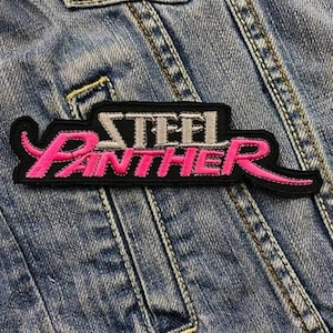 Può includere: Una toppa ricamata nera e rosa con la scritta "STEEL PANTHER" in un font stilizzato. La toppa è rettangolare con un bordo nero ed è applicata su una giacca di jeans.