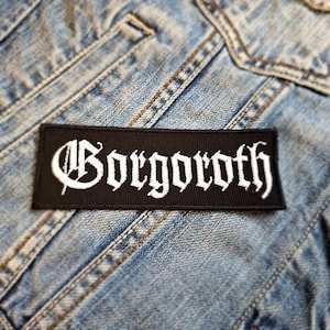 May include: A black rectangular patch featuring the word "Gorgoroth" in white gothic script. The patch is sewn onto a light blue denim jacket, with visible stitching and pocket details.