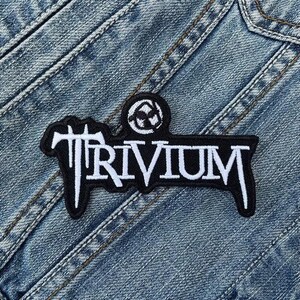 Könnte beinhalten: Ein schwarz-weißer Aufnäher mit dem Wort "TRIVIUM" in einer stilisierten Schriftart. Der Aufnäher hat einen schwarzen Rand und ist auf eine Jeansjacke genäht. Das Patch hat auch ein kleines kreisförmiges Design über dem Wort.