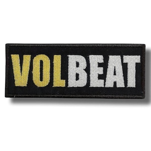 May include: A rectangular black patch with the word "VOLBEAT" embroidered in yellow and white thread. The word "VOL" is in yellow, and "BEAT" is in white. The patch has a black border and is likely for clothing or accessories.