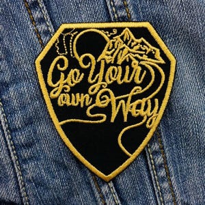Könnte beinhalten: Ein schwarz-goldener, gestickter Aufnäher mit dem Schriftzug "Go Your Own Way" in einem stilisierten Schriftzug. Der Aufnäher zeigt ein Bergketten-Design und ist vor einem Denim-Hintergrund angebracht.