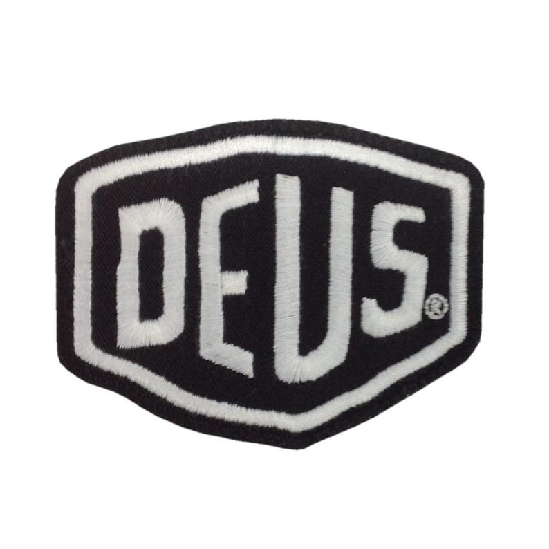 May include: Black and white embroidered patch with the word "DEUS" in bold, stylized letters. The patch has a unique hexagonal shape with a white border. The design is simple and eye-catching, suitable for apparel or accessories.