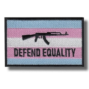 Può includere: Toppa ricamata con i colori della bandiera del pride transgender: azzurro, rosa e bianco. Una silhouette nera di un fucile è centrata sulla toppa. Le parole "DEFEND EQUALITY" sono stampate in nero.
