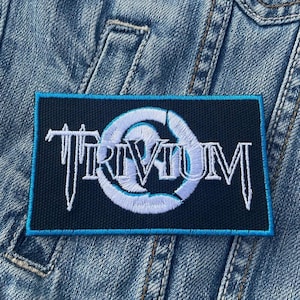 May include: A rectangular patch featuring the word "TRIVIUM" in white, stylized lettering. The text is set against a navy blue background, with a light blue border. The patch is attached to a denim jacket.