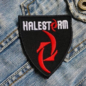Puede incluir: Parche bordado negro con la palabra "HALESTORM" en blanco y un diseño rojo. El parche tiene forma de escudo y está sobre una chaqueta vaquera.