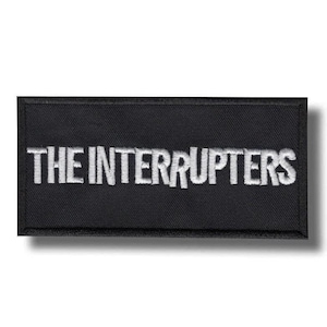 May include: A black rectangular patch featuring the embroidered words "THE INTERRUPTERS" in white. The patch is designed for application on clothing or accessories, with a simple, clear aesthetic.