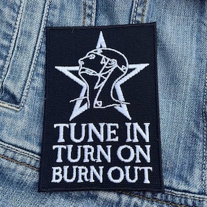 Könnte beinhalten: Schwarzer rechteckiger Aufnäher mit weißem Stern und Profilillustration. Der Aufnäher trägt den weißen Schriftzug "TUNE IN TURN ON BURN OUT". Der Aufnäher befindet sich auf einem Jeansstoff-Hintergrund.