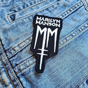 Puede incluir: Un parche en blanco y negro con el texto "MARILYN MANSON" y "MM" sobre un fondo de mezclilla. El parche tiene un diseño único con una cruz en la parte inferior, ideal para ropa o accesorios.