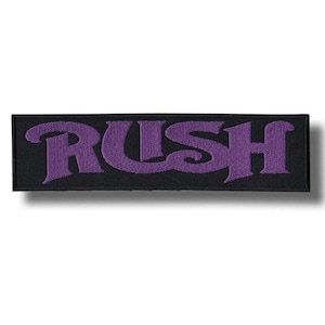 Pode incluir: Patch retangular preto com a palavra "RUSH" bordada em roxo, com letras estilizadas. O patch tem uma borda preta e provavelmente foi projetado para ser costurado ou passado a ferro em roupas ou acessórios. O patch é retangular.