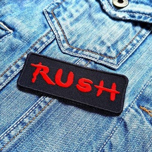 May include: A black rectangular patch with the word "RUSH" embroidered in red thread. The patch is sewn onto a light blue denim jacket, with visible stitching and a button.