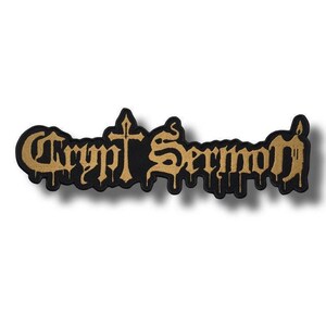Könnte beinhalten: Schwarz-goldener, gestickter Aufnäher mit dem Schriftzug "Crypt Sermon" in gotischer Schrift. Der Aufnäher hat ein Kreuz und einen Tropfeffekt, der eine Vintage-Ästhetik erzeugt.