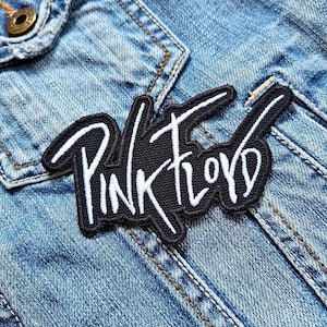 Puede incluir: Un parche bordado negro con las palabras "Pink Floyd" en letras blancas. El parche está cosido en una chaqueta vaquera azul claro. El parche tiene un borde blanco.