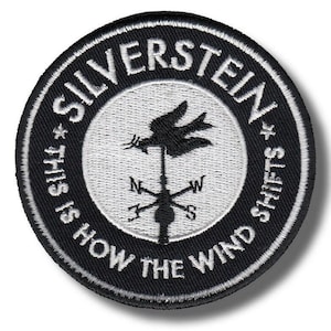 May include: Circular black and white embroidered patch with the text "SILVERSTEIN" and "THIS IS HOW THE WIND SHIFTS". The center features a weather vane design with a bird and compass points.