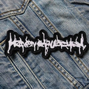 Può includere: Una toppa ricamata in bianco e nero con la scritta "Heavy Love" in un carattere gotico stilizzato. La toppa rettangolare ha un bordo nero ed è posta su uno sfondo di denim blu.