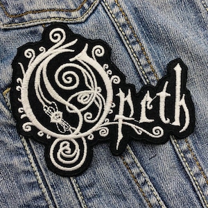 May include: Black and white embroidered patch featuring the word "Opeth" in a gothic font. The patch has a decorative design with swirls and a stylized "O" in the center. The patch is sewn onto a denim jacket.