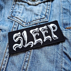 Puede incluir: Un parche rectangular negro con la palabra "SLEEP" bordada en blanco, con letras góticas estilizadas. El parche está colocado sobre una chaqueta vaquera azul descolorida, con costuras visibles y un bolsillo.
