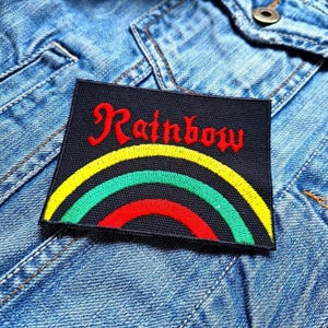 May include: A black rectangular patch with the word "Rainbow" in red script above a yellow, green, and red rainbow design. The patch is sewn onto a blue denim jacket.