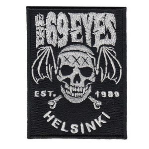 Könnte beinhalten: Schwarzer rechteckiger Aufnäher mit weiß gesticktem Text und Totenkopf-Design. Der Text lautet "THE 69 EYES", "EST. 1989" und "HELSINKI". Der Totenkopf hat Fledermausflügel und gekreuzte Knochen.