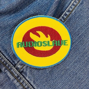 Puede incluir: Un parche circular con la palabra "AUDIOSLAVE" en letras verdes. El parche tiene un fondo amarillo con un diseño rojo en forma de llama en el centro y un borde azul. El parche está cosido sobre una tela vaquera.