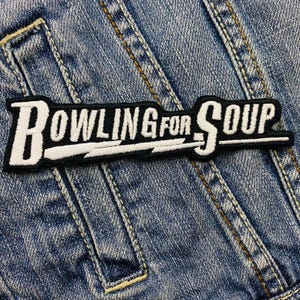 Könnte beinhalten: Gestickter Aufnäher mit dem Text "BOWLING FOR SOUP" in Weiß, umrandet in Schwarz, auf schwarzem Grund. Der Aufnäher ist auf einer Jeansjacke befestigt. Der Text ist mit einem Blitz gestaltet.