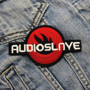 Puede incluir: Parche bordado con el texto "AUDIOSLAVE" en blanco sobre fondo negro. El parche presenta un círculo rojo con un diseño de llama negra. El parche está colocado sobre un fondo de mezclilla.