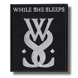May include: Black square patch with the text "WHILE SHE SLEEPS" at the top. Below the text is a stylized silver emblem featuring intertwined "W" and "S" letters. Two small silver diamond shapes flank the emblem.