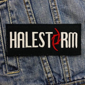 Puede incluir: Parche rectangular negro con la palabra "HALESTORM" bordada en letras mayúsculas blancas. Un diseño rojo estilizado está entre la "T" y la "R". El parche está sobre una chaqueta vaquera.