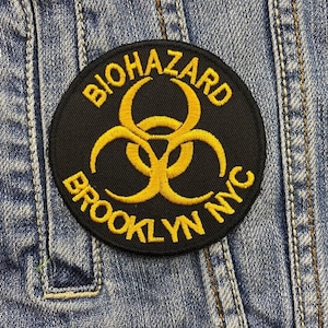 May include: A round black patch with yellow embroidered text and a biohazard symbol. The text reads "BIOHAZARD BROOKLYN NYC". The patch is sewn onto a blue denim jacket.