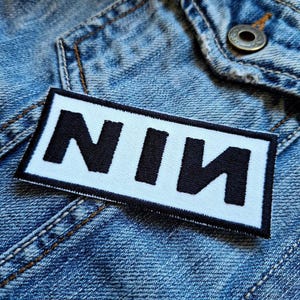 Könnte beinhalten: Ein rechteckiger Aufnäher mit den Buchstaben "NIN" in fetter schwarzer Schrift auf weißem Hintergrund. Der Aufnäher ist mit schwarzen Nähten eingefasst und auf einer Jeansjacke befestigt.