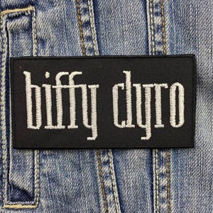 Pode incluir: Um patch retangular preto com o texto bordado branco "biffy clyro" está afixado a uma jaqueta jeans. O patch está centralizado e o texto está em uma fonte estilizada. A jaqueta jeans tem costuras e bolsos visíveis.