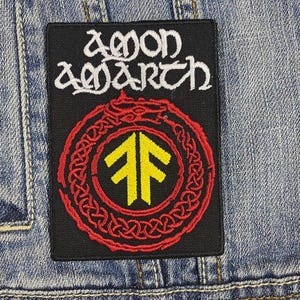 Può includere: Toppa rettangolare nera con le parole "Amon Amarth" in bianco, sopra un disegno circolare rosso e giallo. La toppa è cucita su una giacca di jeans.