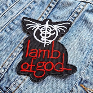 May include: Black patch with the words "lamb of god" in red lettering. The patch has a white emblem at the top. The patch is on a denim jacket.