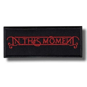 Può includere: Toppa rettangolare nera con la scritta ricamata "IN THIS MOMENT" in rosso, con decorazioni. La toppa è probabilmente per abbigliamento o accessori.