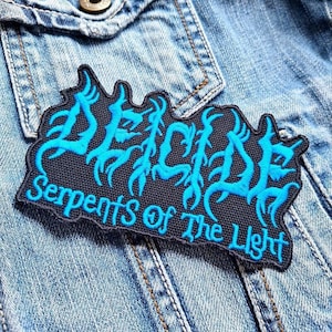 May include: A black embroidered patch featuring the word "DECADE" in turquoise, stylized lettering, and the text "Serpents of The Light". The patch is sewn onto a light blue denim jacket.