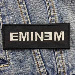 May include: A black rectangular patch with the word "EMINEM" embroidered in white, set against a blue denim background. The patch is likely designed to be sewn or ironed onto clothing or accessories.