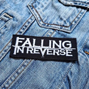 May include: A black rectangular patch embroidered with the words "FALLING IN REVERSE" in white. The patch is attached to a light blue denim jacket, showing visible stitching and pocket details.