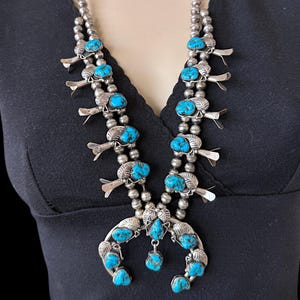 May include: A silver squash blossom necklace with turquoise stones. The necklace features a double strand of silver beads, with turquoise stones set in silver blossoms and a crescent-shaped pendant. The necklace is displayed on a mannequin.