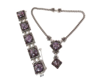 May include: A silver necklace and bracelet set featuring purple iridescent glass stones. The necklace has a pendant in a teardrop shape. The bracelet has a chain link design.