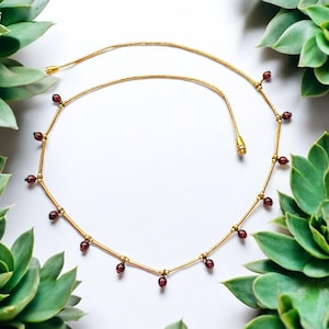 Könnte beinhalten: Eine zarte goldene Halskette mit kleinen, runden, roten Perlen, die gleichmäßig verteilt sind. Die Halskette wird vor einem weißen Hintergrund präsentiert, umgeben von grünen Sukkulenten. Die Halskette hat ein schlichtes, elegantes Design.