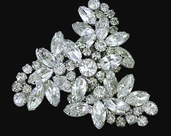 May include: A sparkling, silver-toned brooch with a floral design. The brooch features a cluster of clear, faceted crystals in various shapes and sizes, including marquise and round cuts. The crystals are set against a dark background.