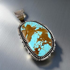 May include: Oval turquoise pendant with a silver setting. The pendant features a large, polished turquoise stone with brown and blue hues. The silver setting has a decorative rope-like border and a bail for a chain.