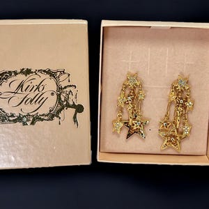 May include: A pair of gold-tone star-shaped dangle earrings with clear crystals. The earrings are in a box with the words "Kirk's Folly" printed on the lid.