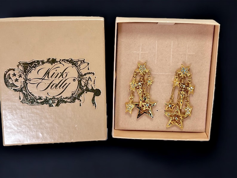 May include: A pair of gold-tone star-shaped dangle earrings with clear crystals. The earrings are in a box with the words "Kirk's Folly" printed on the lid.