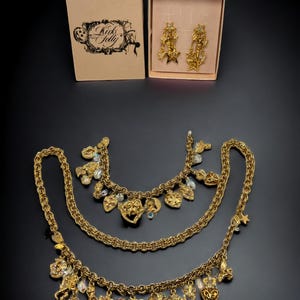 May include: A gold-tone necklace set with multiple charms, including hearts, stars, and other decorative elements. The set also includes a pair of matching earrings. The jewelry is packaged in two pink boxes with the text "Kink & Folly" on one of the boxes.